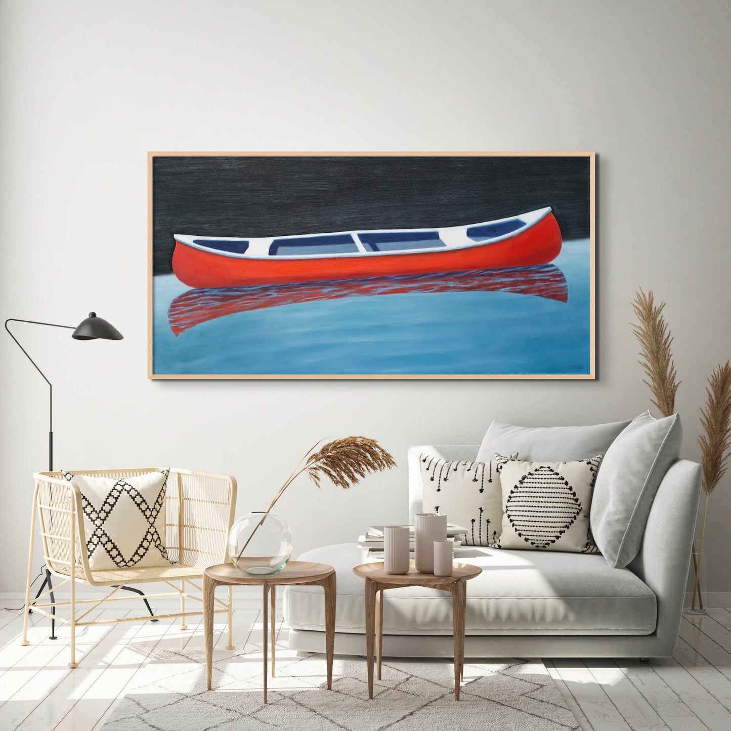 Canoe painting of a small red boat by Canadian artist Catherine McKinnon. The red canoe is floating on almost still blue water in the foreground and the background is pitch black. The giclee print is framed in light colored wood and is mounted on a light gray wall above a stylish contemporary chaise and living room set-up.