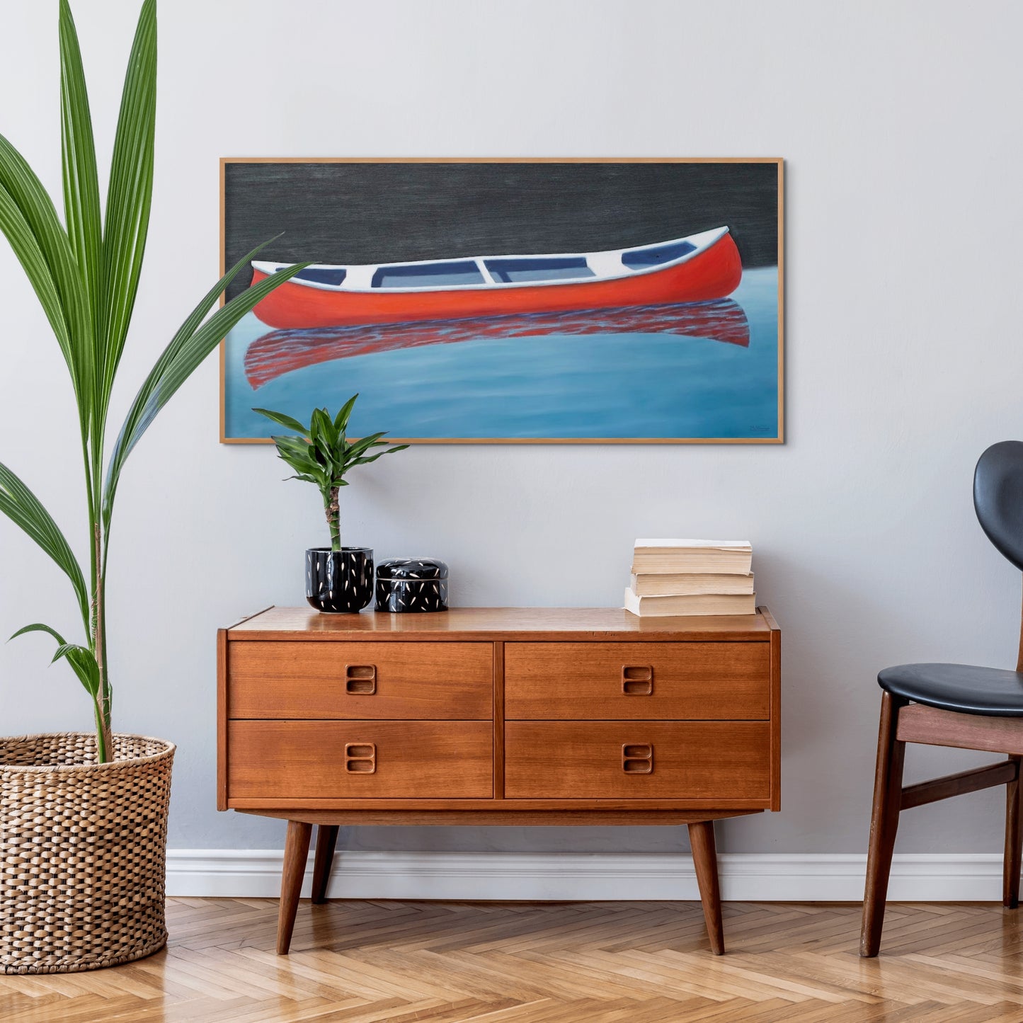 Canoe painting of a small red boat by Canadian artist Catherine McKinnon. The red canoe is floating on almost still blue water in the foreground and the background is pitch black. The giclee print is framed in cherry wood. It is mounted on a light gray wall above a mid-century modern cherry wood sideboard.