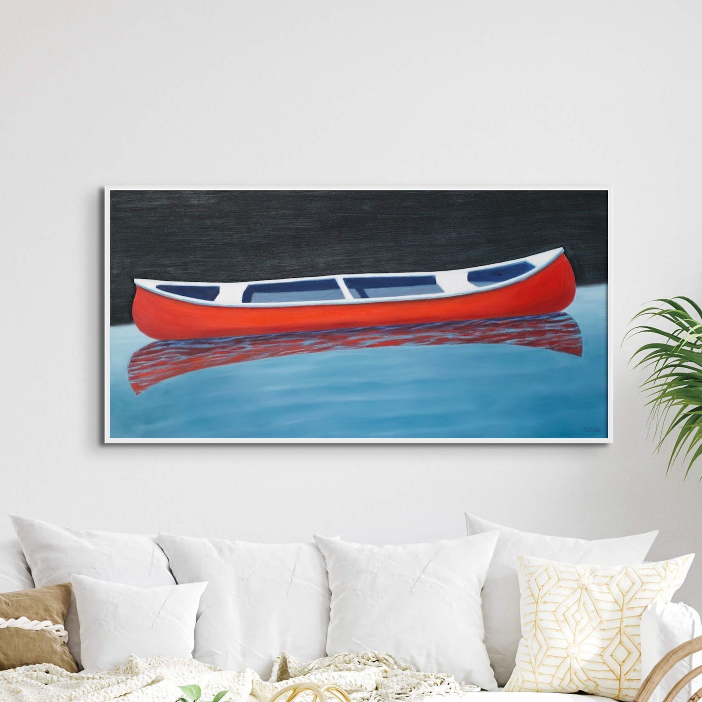A painting of a red canoe by Canadian artist Catherine McKinnon. The small red boat is floating on almost still blue water in the foreground and the background is pitch black. The giclee print is framed in white. It is mounted on a light gray wall above a stylish, contemporary couch with many white cushions.