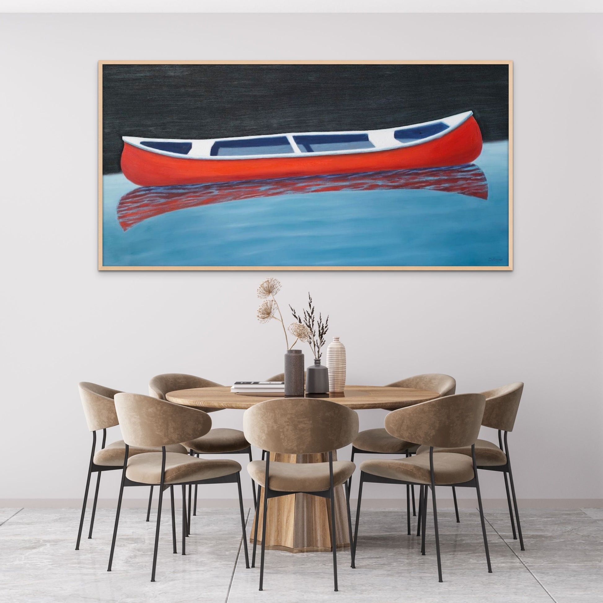 A painting of a red canoe by Canadian artist Catherine McKinnon. The small red boat is floating on almost still blue water in the foreground and the background is pitch black. The giclee print is framed in light colored wood. It is mounted on a light gray wall above a stylish, luxury, contemporary dining room table and seven chairs.