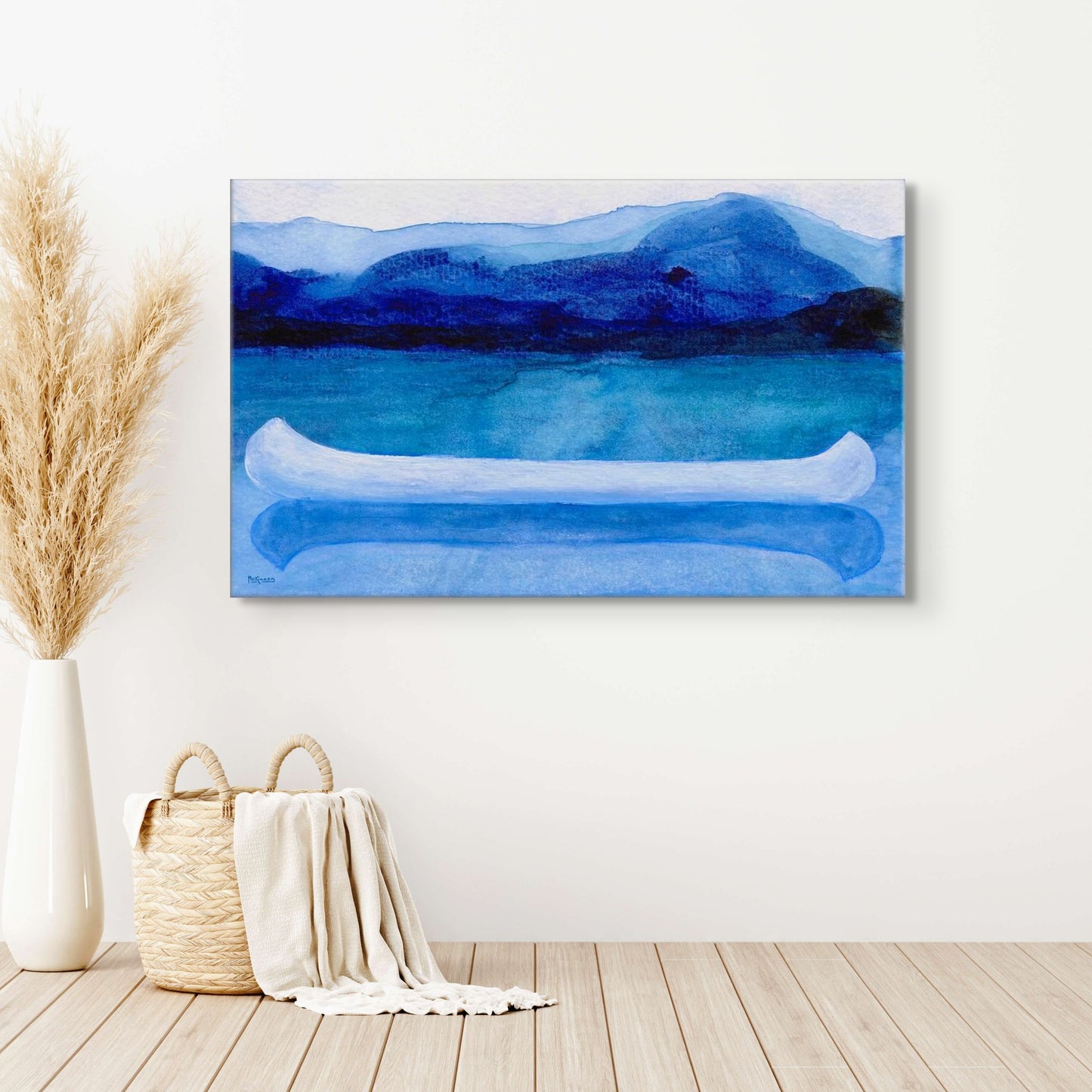 A work of canoeing art by Canadian artist Catherine McKinnon. The watercolor depicts a small white boat, a "canoe", on blue water with a dark blue distant shore and lighter blue distant moutains. The giclee print is frameless and is mounted on a white wall above a beachy wood floor, straw beach tote, draped blanket and white vase with tall wheat-colored fronds.