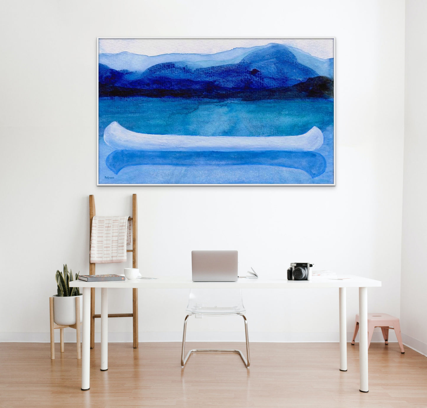 A work of canoeing art by Canadian artist Catherine McKinnon. The watercolor depicts a small white boat, a "canoe", on blue water with a dark blue distant shore and lighter blue distant moutains. The giclee print is framed in white and is mounted on a white wall above a white, stylish, contemporary desk and chair..