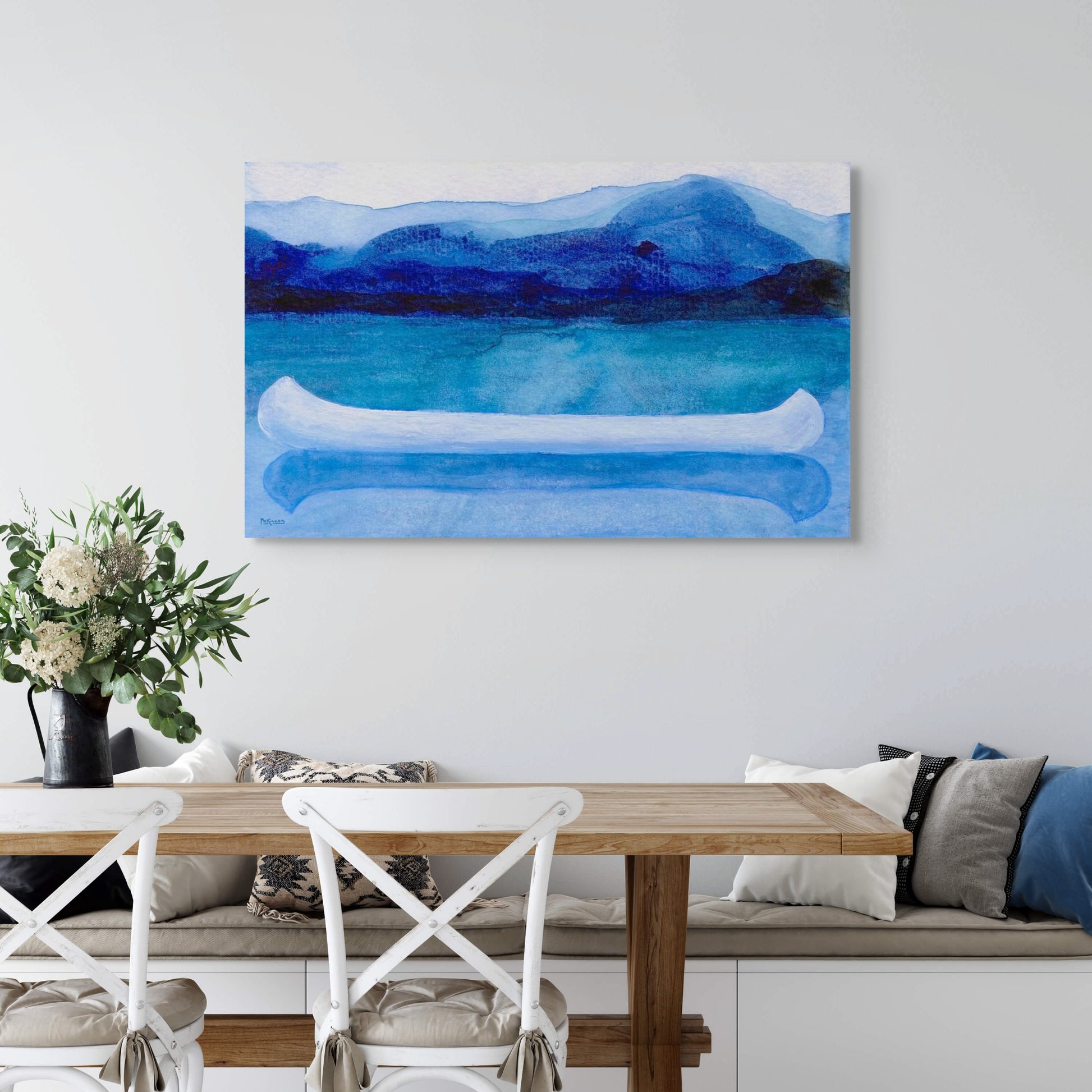 A work of canoeing art by Canadian artist Catherine McKinnon. The watercolor depicts a small white boat, a "canoe", on blue water with a dark blue distant shore and lighter blue distant moutains. The giclee print is frameless and is mounted on a white wall above a modern farmhouse table.