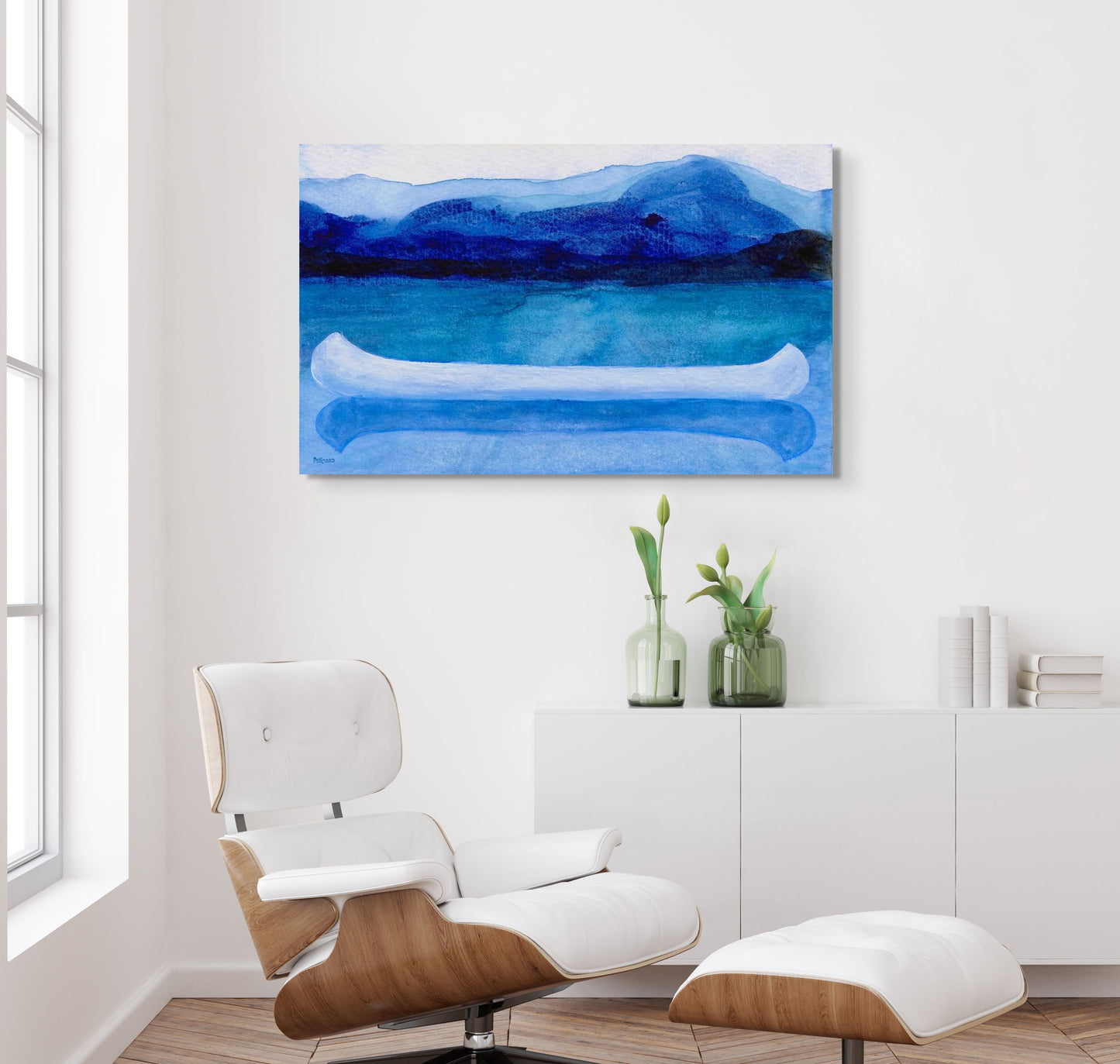 A work of canoeing art by Canadian artist Catherine McKinnon. The watercolor depicts a small white boat, a "canoe", on blue water with a dark blue distant shore and lighter blue distant moutains. The giclee print is frameless and is mounted on a white wall above a stylish, contemporary white lounging chair with natural wood structure.