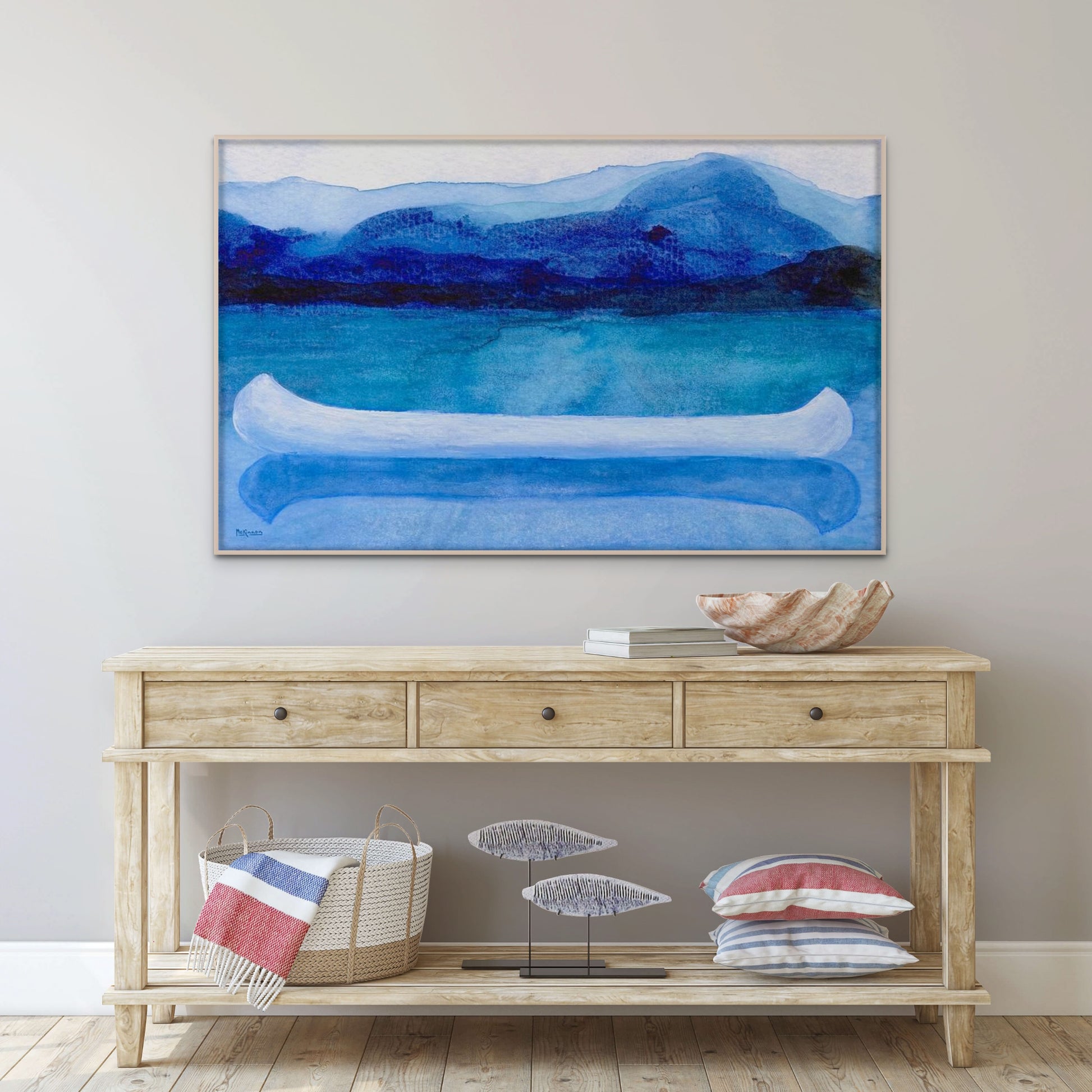 A work of canoeing art by Canadian artist Catherine McKinnon. The watercolor depicts a small white boat, a "canoe", on blue water with a dark blue distant shore and lighter blue distant moutains. The giclee print is framed in white and is mounted on a light gray wall above a beachy, light colored wood sideboard and beach house decor items.