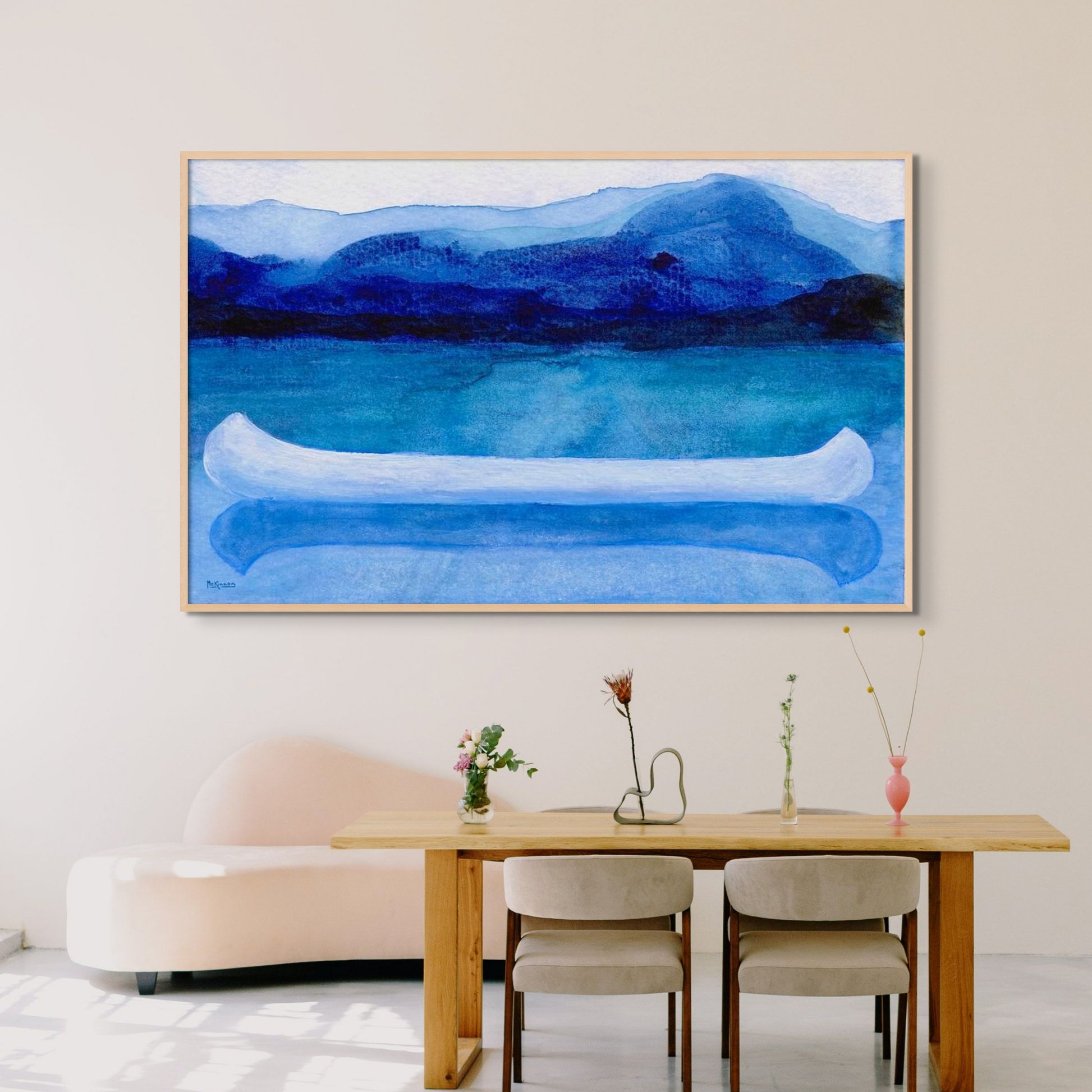 A work of canoeing art by Canadian artist Catherine McKinnon. The watercolor depicts a small white boat, a "canoe", on blue water with a dark blue distant shore and lighter blue distant moutains. The giclee print is framed in light natural wood and is mounted on a light gray wall above a beachy, light colored wood dining table and modern cream-colored settee.